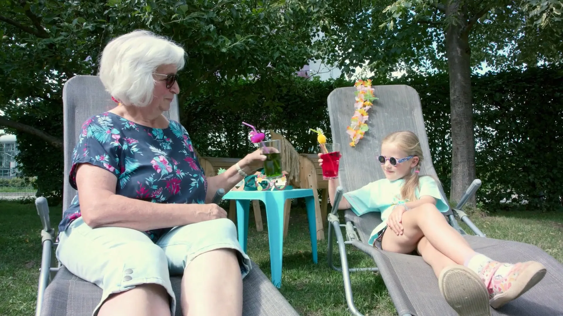 Seniorin und Mädchen mit Sonnenbrillen und bunten Cocktails auf Liegestühlen im Garten, Standbild aus dem Imagefilm für die Volkssolidarität Gera